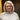 Becky Baker stands in front of a wooden door in a yellow wall. She is wearing a beige One Sweater and has short wavy hair and glasses.